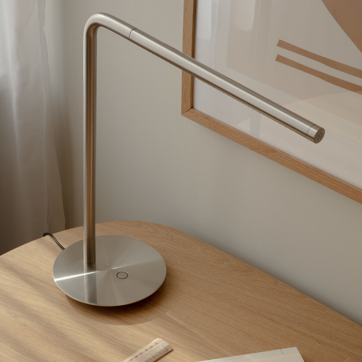 Moderne, minimalistische Tischleuchte aus gebürstetem Metall steht auf einem hellen Holztisch neben Schreibutensilien, einem Lineal und einer Notizmappe, im Hintergrund helle Wanddekoration und Tageslicht von einem Fenster.