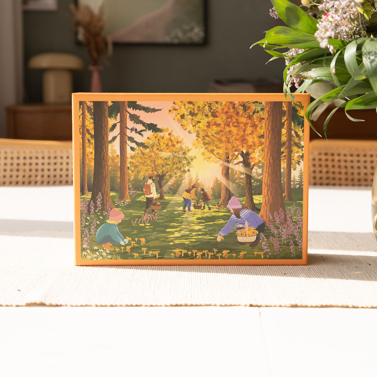 Das Bild zeigt einen Karton des 1000-Teile Puzzles »Waldschätze« auf einem hellen Tisch. Das Puzzle-Motiv zeigt eine gemalte Waldszene im Herbst mit Menschen, darunter Kinder, die Pilze und Waldschätze sammeln. Sonnenstrahlen fallen durch die bunten Laubbäume. Im Vordergrund steht ein Blumenstrauß.