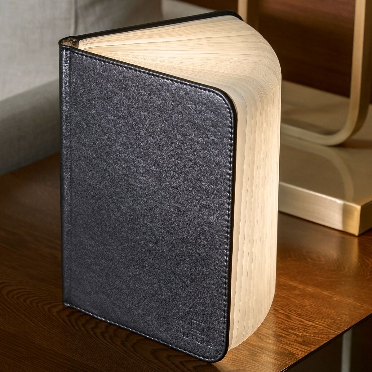 Ein Smart Booklight in schwarzem Lederdesign, das wie ein aufgeschlagenes Buch aussieht. Es steht auf einem Holztisch vor einer Couch und bietet eine warme LED-Innenbeleuchtung.