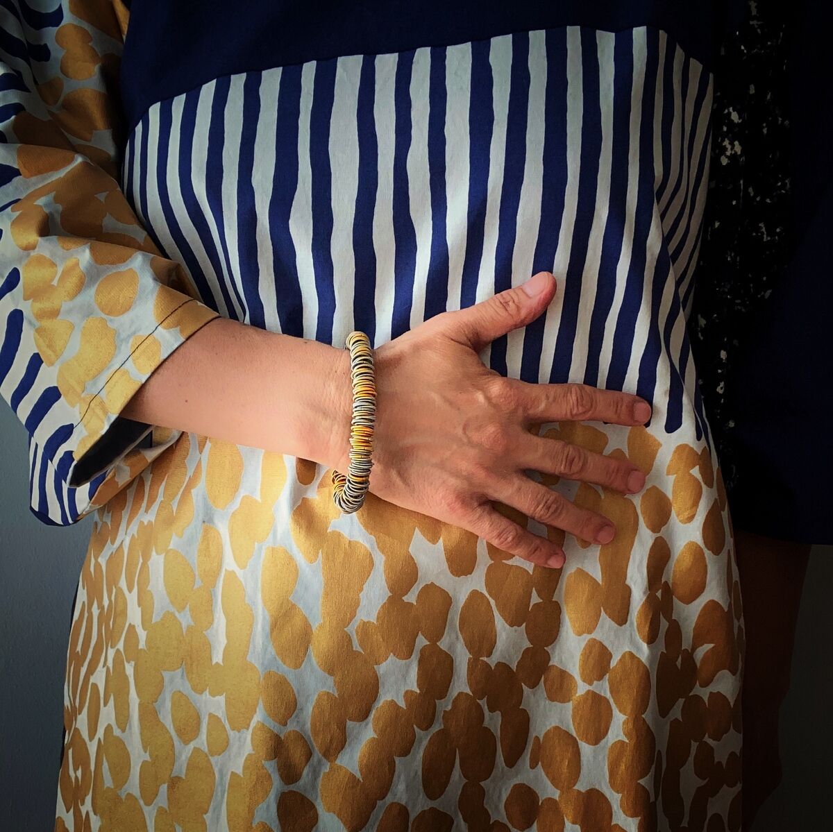 Eine Person trägt ein Armband am Handgelenk. Das Armband ist gelb und braun. Sie trägt ein Kleid mit blau-weißen und gelb-bräunlichen Mustern.
