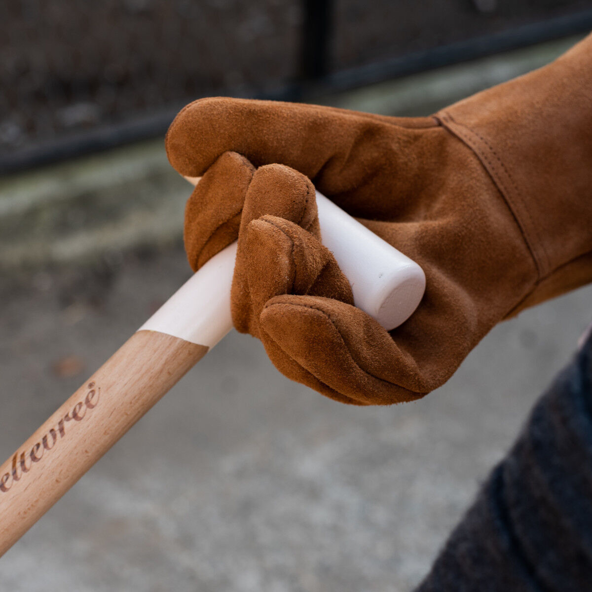 Ein Paar braune Handschuhe aus Rindleder hält einen Holzstiel. Die Handschuhe sehen robust und bequem aus, ideal für Arbeiten im Freien oder handwerkliche Tätigkeiten.