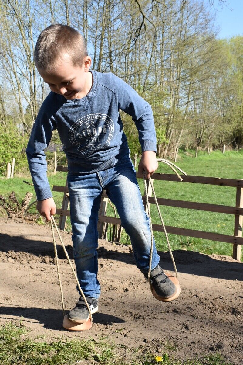 Ein Junge spielt draußen mit Shaddy Spielhufen. Er steht auf den hölzernen Hufen mit Seilen und hält die Seile in den Händen. Im Hintergrund sind Bäume und ein Gartenzaun zu sehen.