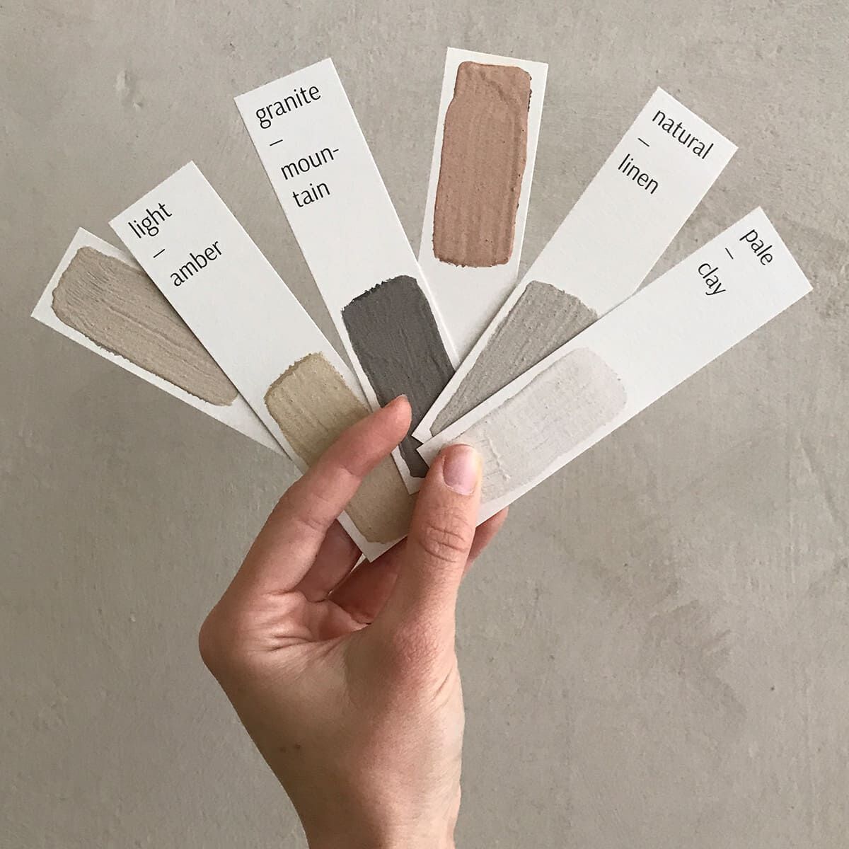 Eine Hand hält einen Farbfächer mit fünf verschiedenen Lehmfarben. Die Farbnuancen variieren von hellem Amber über Granit und Mountain bis hin zu Naturleinen und hellem Ton. Der Hintergrund ist neutral und schlicht, was die Farben in den Vordergrund rückt.