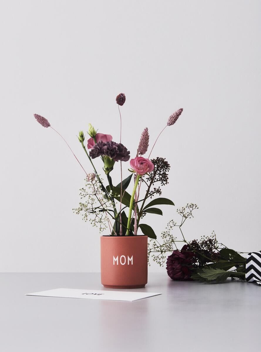 Ein terrakottafarbener Becher mit der Aufschrift 'MOM', gefüllt mit verschiedenen rosa und grünen Blumen. Im Hintergrund befindet sich ein weiterer Blumenstrauß und eine weiße Karte auf einer grauen Oberfläche.
