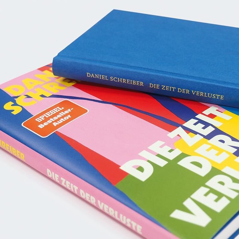 Ein Stapel von Büchern des Titels 'Zeit der Verluste' von Daniel Schreiber. Das obere Buch hat einen blauen Einband, während das untere ein farbenfrohes Cover mit den Farben Blau, Rot, Pink, Gelb und Weiß zeigt. Der Titel und der Autor sind auf dem Buchcover gut sichtbar.