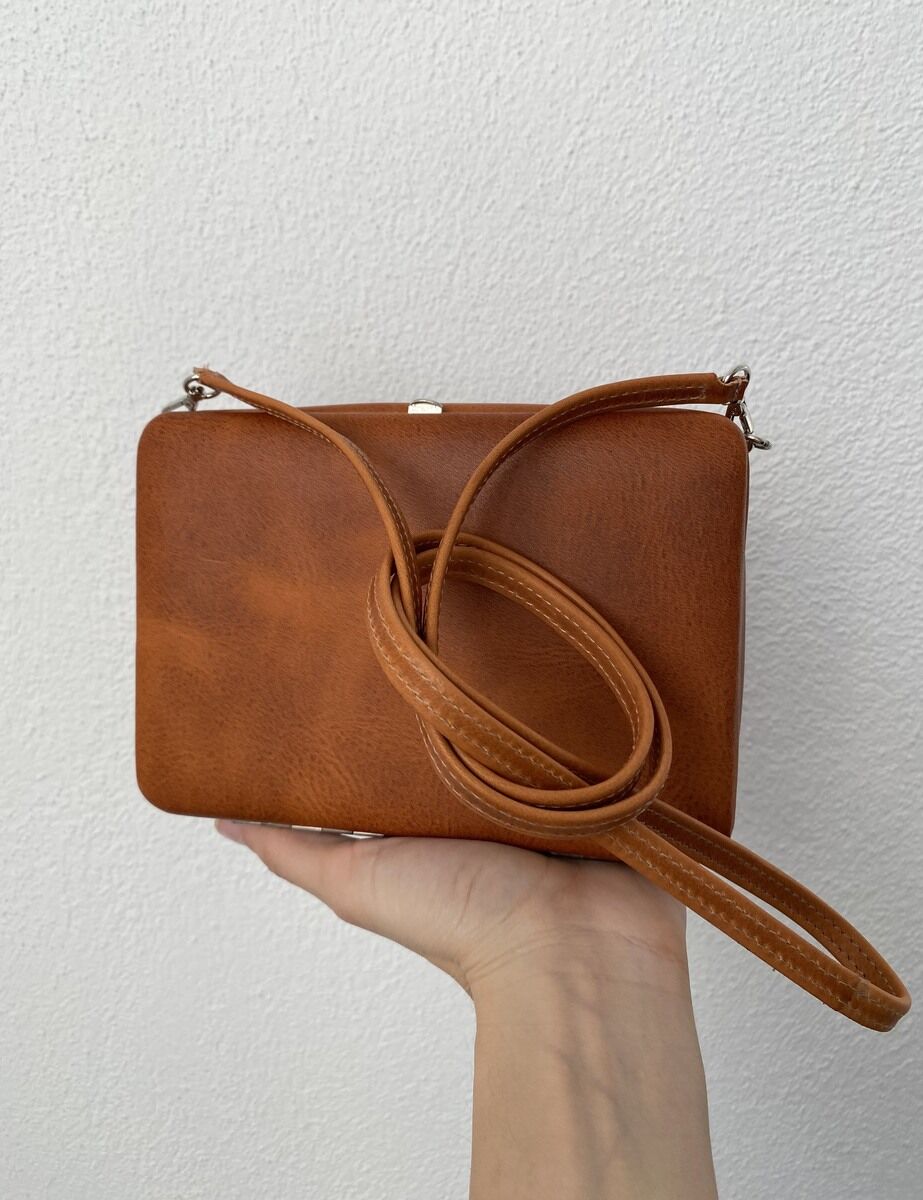 Eine braune Ledertasche mit einem langen Riemen auf einer Hand vor einem hellen Hintergrund.