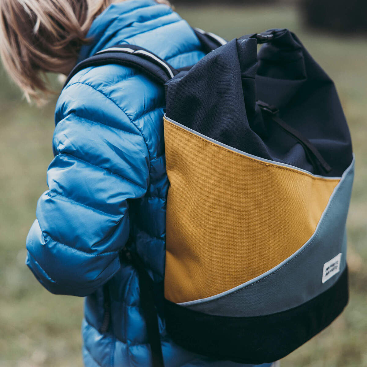 Eine mehrfarbige (Blau, Gelb, Schwarz und Grau) Tasche »Popoyo« als Rucksack getragen von einem Kind in einer blauen Winterjacke. Der Hintergrund zeigt eine natürliche Landschaft mit Gras.