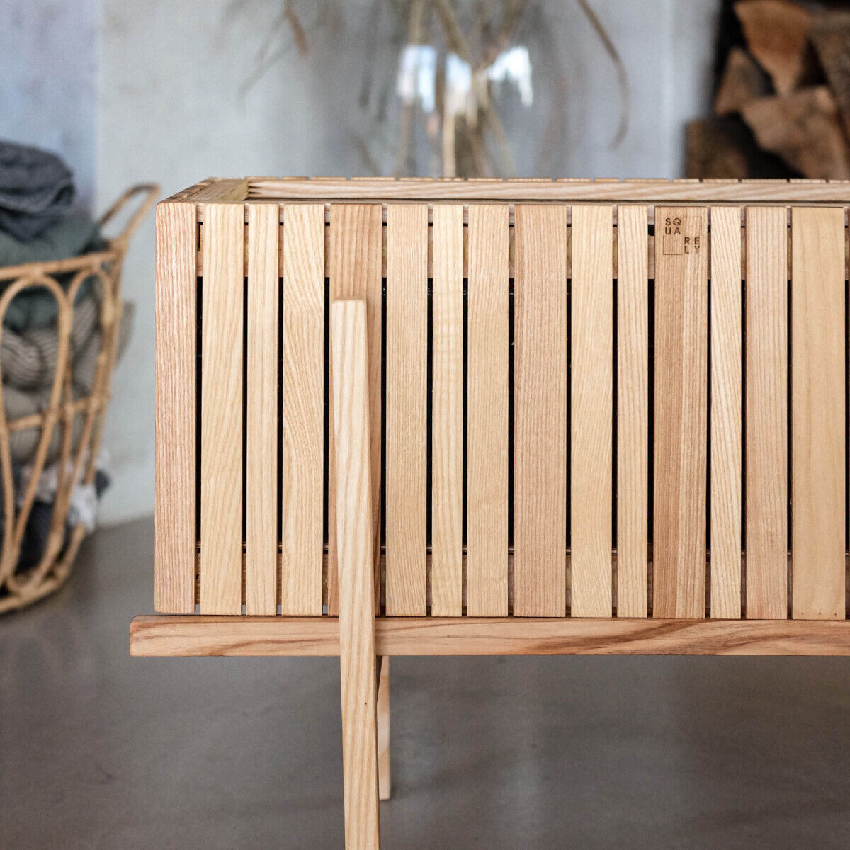 Pflanzgefäß »GrowWIDE« aus Holz mit einem schlichten, modernen Design steht auf einem hellbraunen Holzständer. Im Hintergrund sind unscharf ein geflochtener Korb und ein beige Tuch zu sehen.