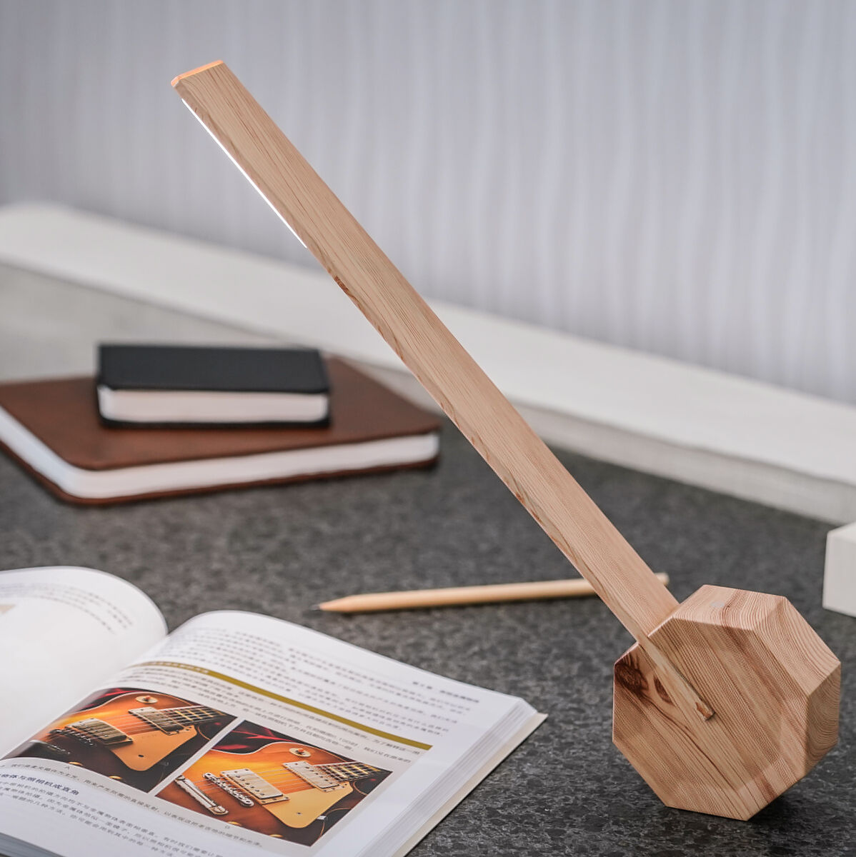 Eine moderne Tischlampe aus Holz in einem minimalistischen Design steht auf einem grauen Tisch. Das Licht der Lampe fällt auf ein aufgeschlagenes Buch. Im Hintergrund befinden sich ein geschlossenes Notizbuch, ein weiteres Buch und eine digitale Uhr mit der Anzeige '10:00'.