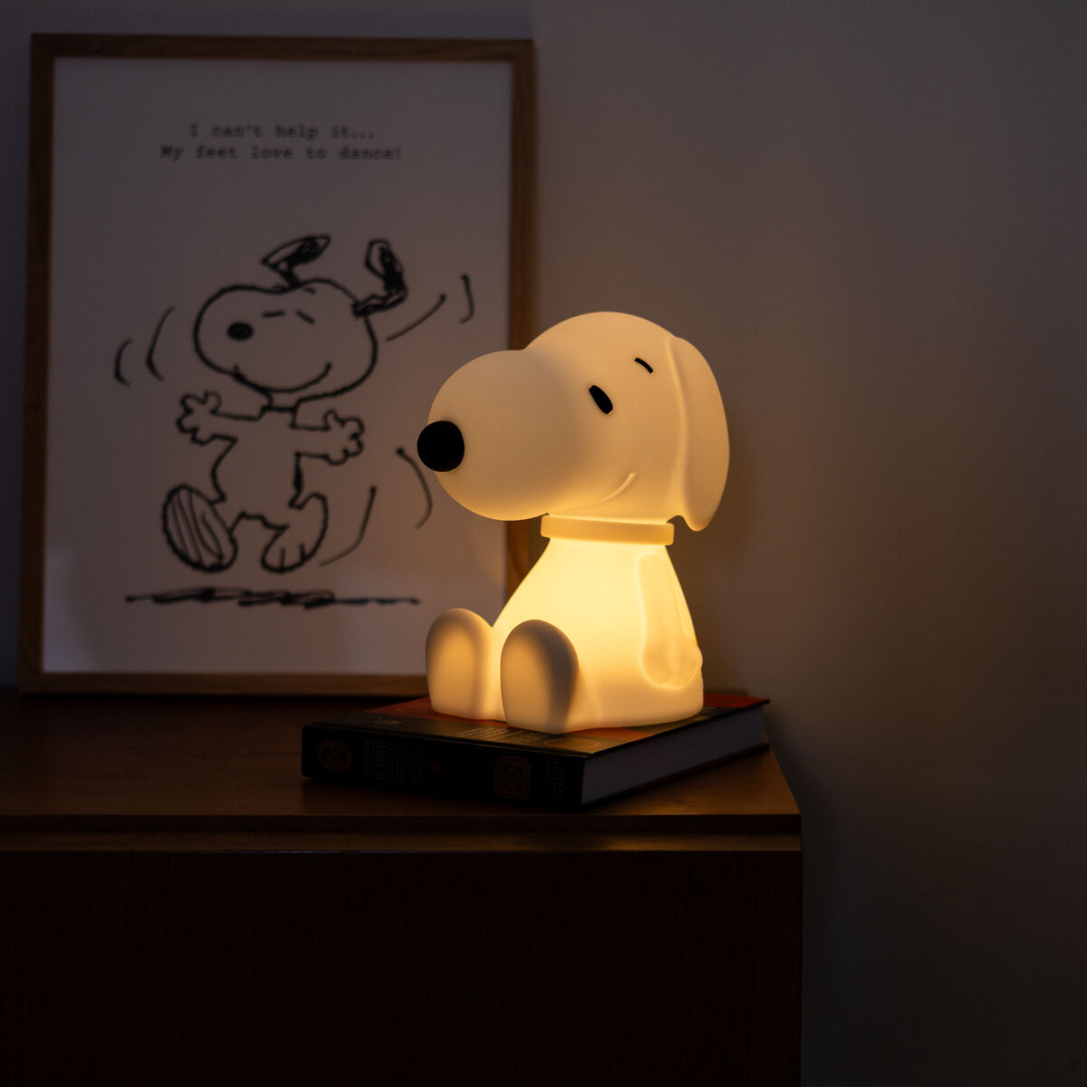 Ein Schneemann-artiges Nachtlicht in Form von Snoopy sitzt auf einem braunen Holztisch. Im Hintergrund befindet sich ein gerahmtes Bild von Snoopy. Das sanfte Licht verleiht dem Raum eine gemütliche Atmosphäre.