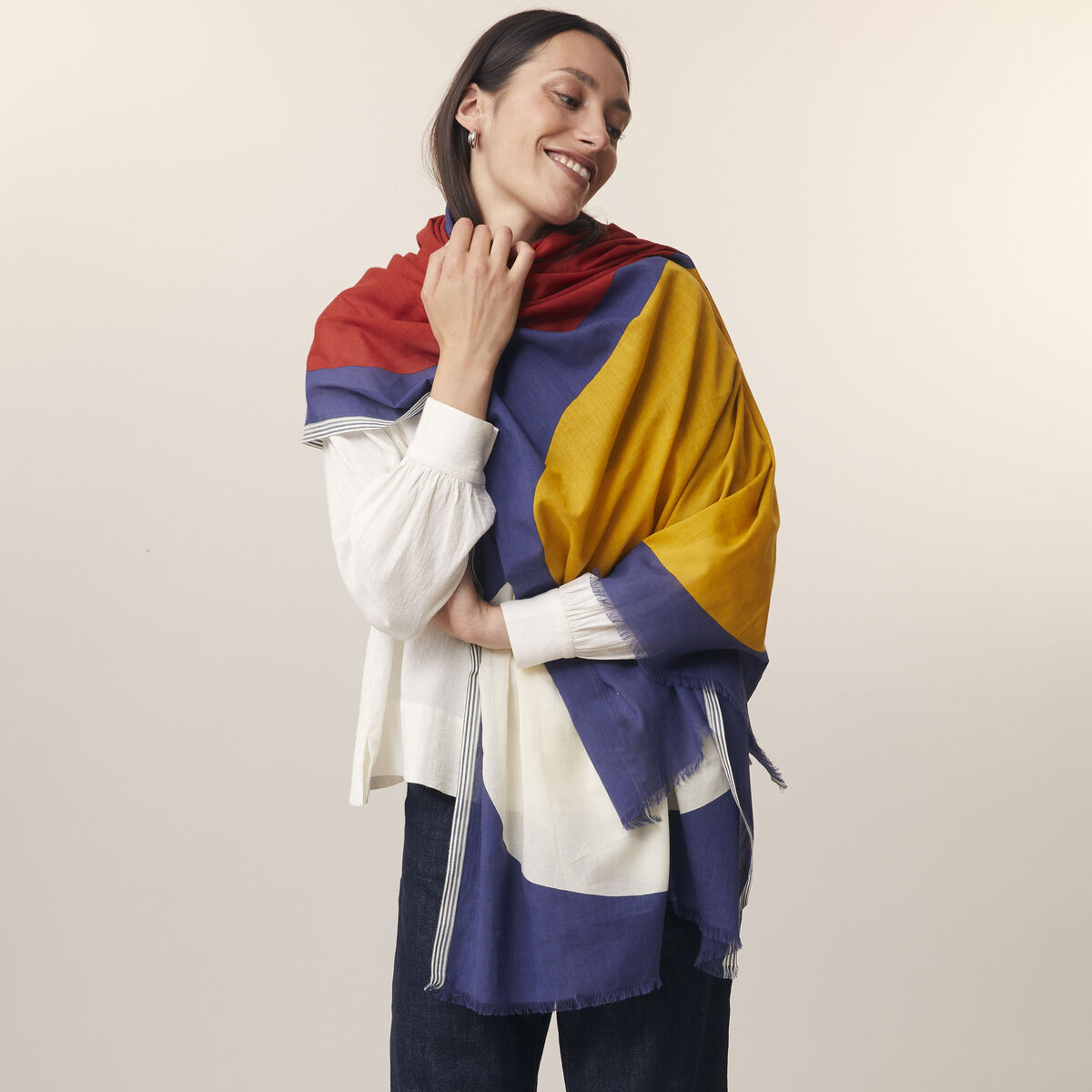 Eine Frau trägt einen bunten Baumwollschal mit roten, blauen und gelben Farbbereichen über ihrer Schulter. Sie ist lässig gekleidet in einem weißen Shirt und dunklen Jeans, steht aufrecht und lächelt sanft in einem neutralen Studioumfeld.