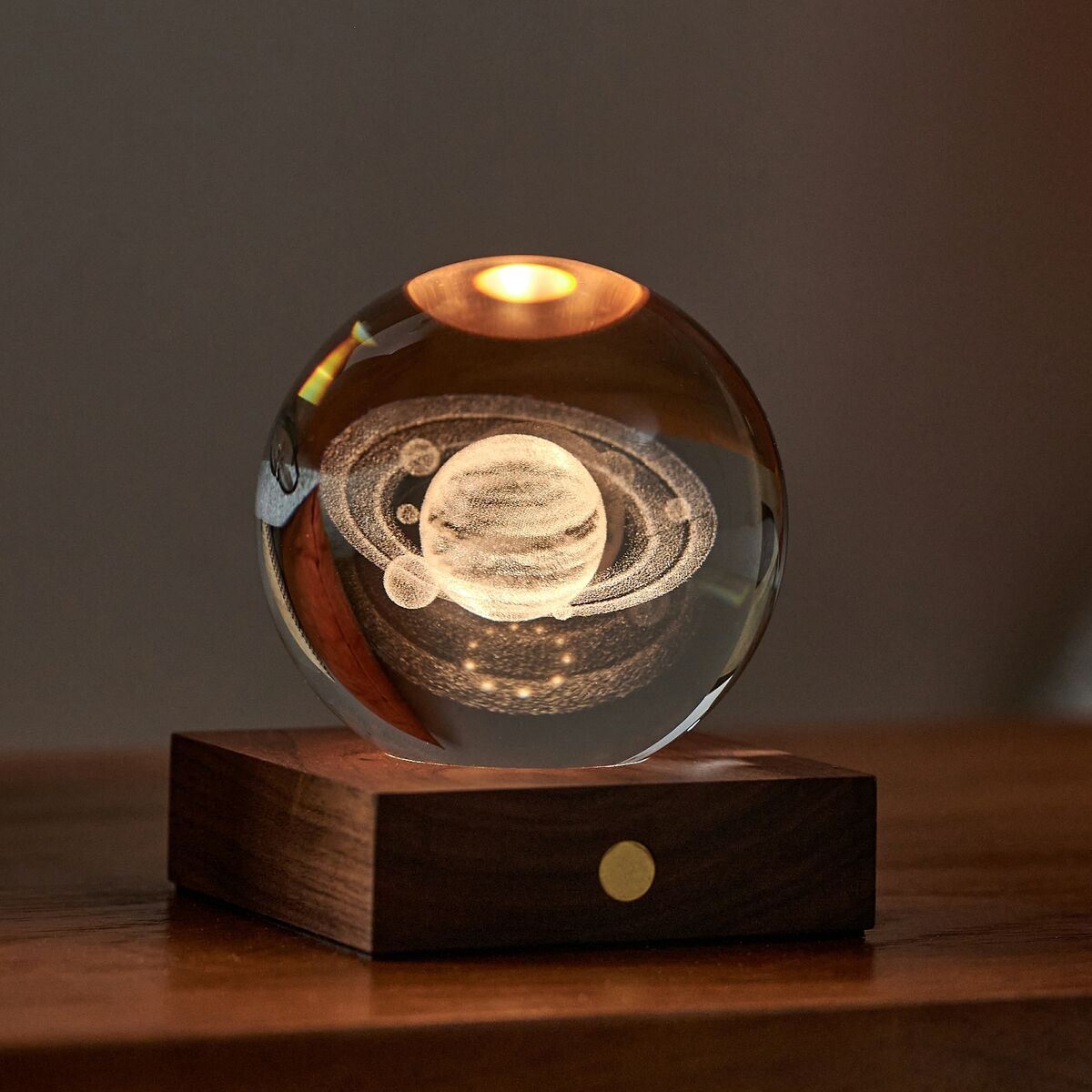 Eine dekorative Kugellampe namens »Amber Light« steht auf einem Holzboden. Sie leuchtet von oben in warmem Bernsteinlicht, in der Glaskugel ist ein 3D-Bild des Planeten Saturns eingraviert. Im Hintergrund befinden sich ein orangefarbenes Kissen und eine stilvoll platzierte Brille.