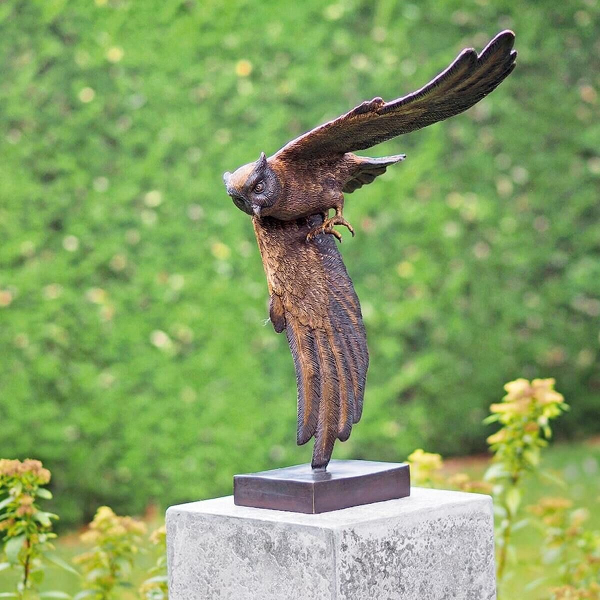 Metallskulptur einer fliegenden Eule, detailreich gestaltet, auf einem Sockel montiert, vor einem Hintergrund aus grünen Büschen.