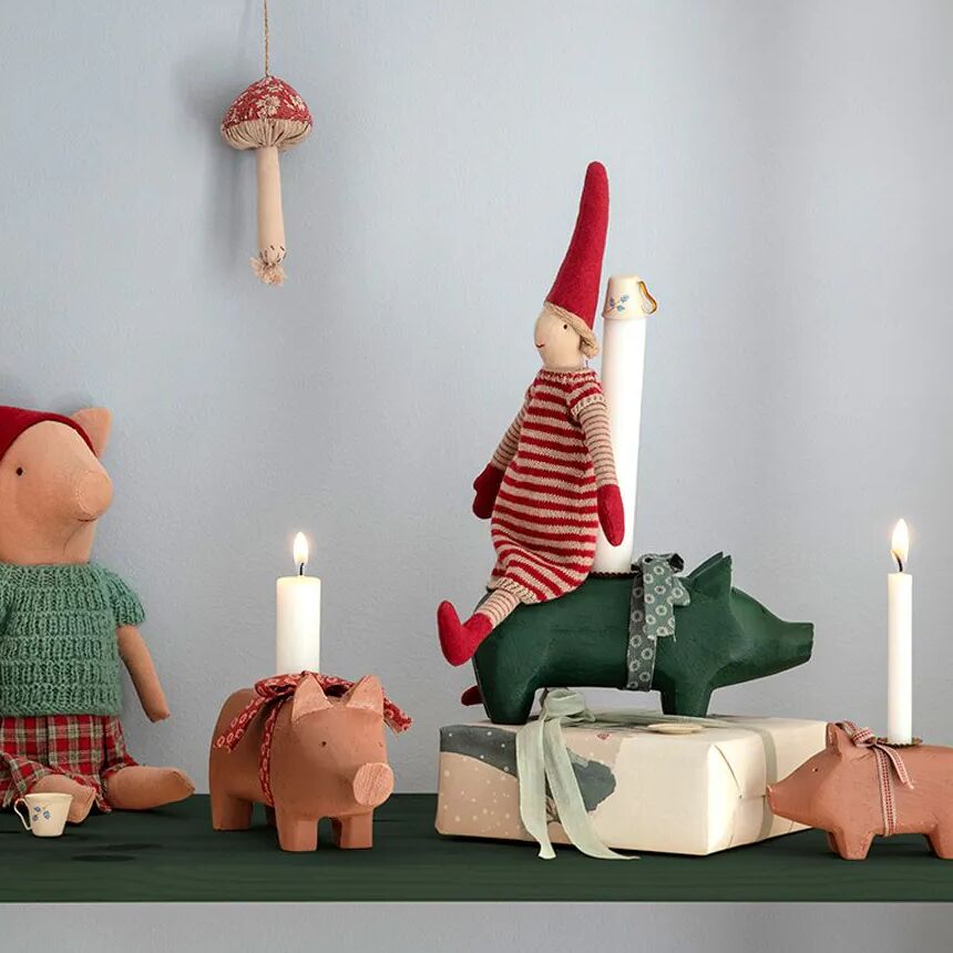 Mittlerer rosa Kerzenhalter in Schwein-Form steht auf einem grünen Regal, flankiert von weiteren Schwein-Kerzenhaltern, verpackten Geschenken mit Schleifen und skandinavischen Stofffiguren in Rot-Weiß, vor hellgrauer Wand mit dekorativem Pilzanhänger. Die Szene vermittelt verspielt-gemütliche Kinderzimmer- oder Festtagsdeko.