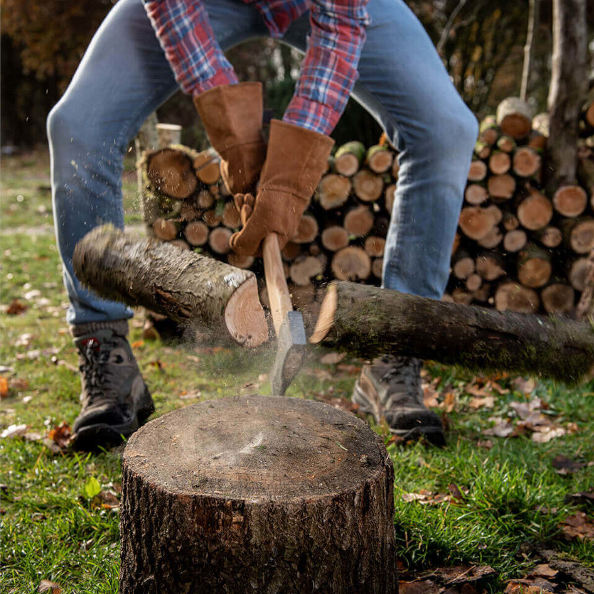 Eine Person in Jeans und einem karierten Hemd hackt mit Hilfe von braunen Rindleder-Handschuhen Holz draußen. Ein Baumstumpf dient als Unterlage, während im Hintergrund gestapeltes Holz zu sehen ist.