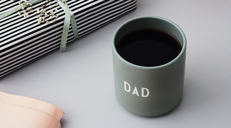 Graue Tasse mit schwarzem Kaffee und weißer Aufschrift 'DAD' neben Geschenk mit schwarz-weiß gestreiftem Papier und grüner Schleife