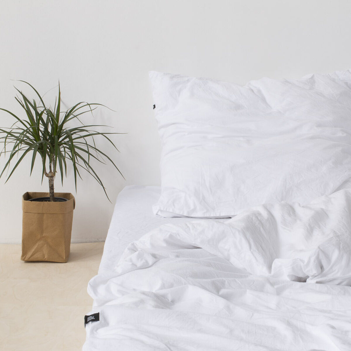 Eine minimalistische Bettwäsche namens „Pure“ in Weiß, arrangiert auf einem Bett mit weißen Kissen und Bezügen. Daneben steht eine grüne Zimmerpflanze in einem braunen Papierübertopf auf dem Boden.