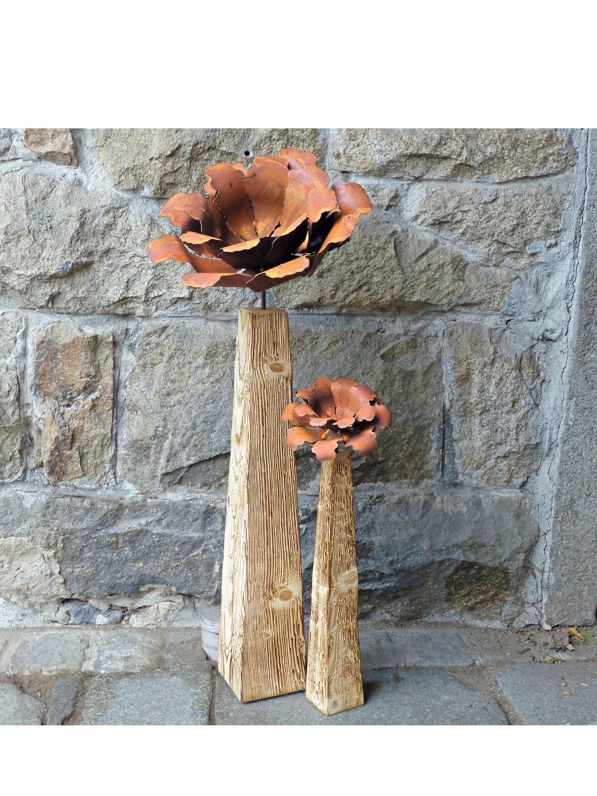 Zwei Gartenstelen mit Kamelienblüten aus Metall vor einer Steinwand. Die Stelen sind aus Holz mit rustikaler Struktur und die Blüten sind in Rostrot und Orange gehalten.