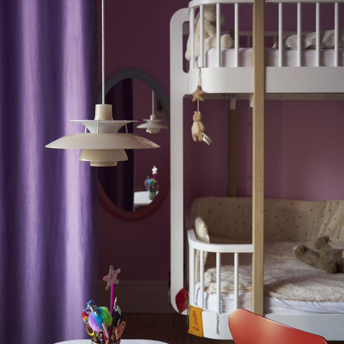 Im Vordergrund hängt die Pendelleuchte »PH 5 320« von Louis Poulsen in opal-beige über einem Kinderschreibtisch mit Bastelutensilien. Im Hintergrund ein Hochbett aus weißem Holz, lila Vorhang, rosafarbene Wand und runde Spiegel.