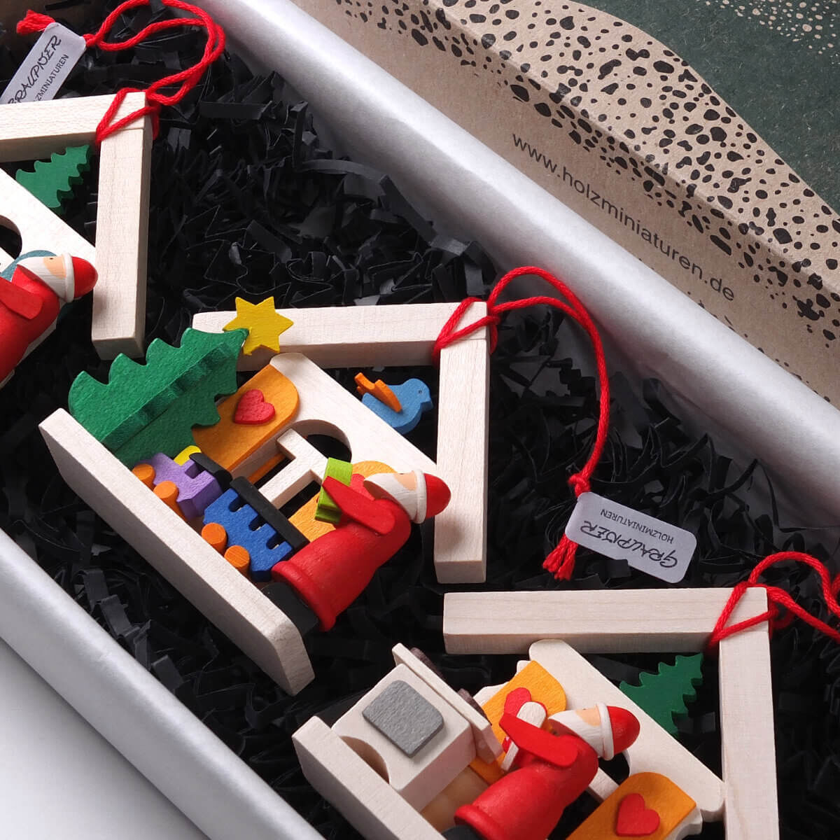 Dreiteiliges Geschenk-Set aus Holz, bestehend aus Weihnachtsmannhäusern mit roten Weihnachtsmännern und Weihnachtselementen, dekoriert mit roten Aufhängern, auf schwarzem Füllmaterial in einer Geschenkschachtel präsentiert.