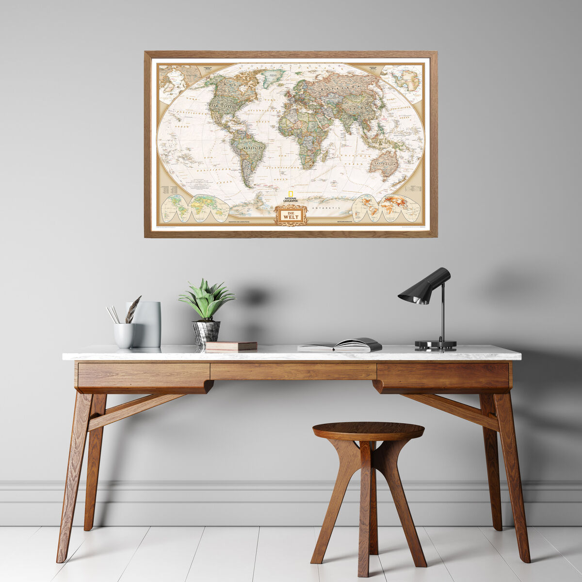 Eine antike Weltkarte von National Geographic, gerahmt und an einer grauen Wand über einem modernen Holzschreibtisch montiert. Der Schreibtisch enthält eine Lampe, einen Blumentopf, Bücher und einen Stiftbehälter.