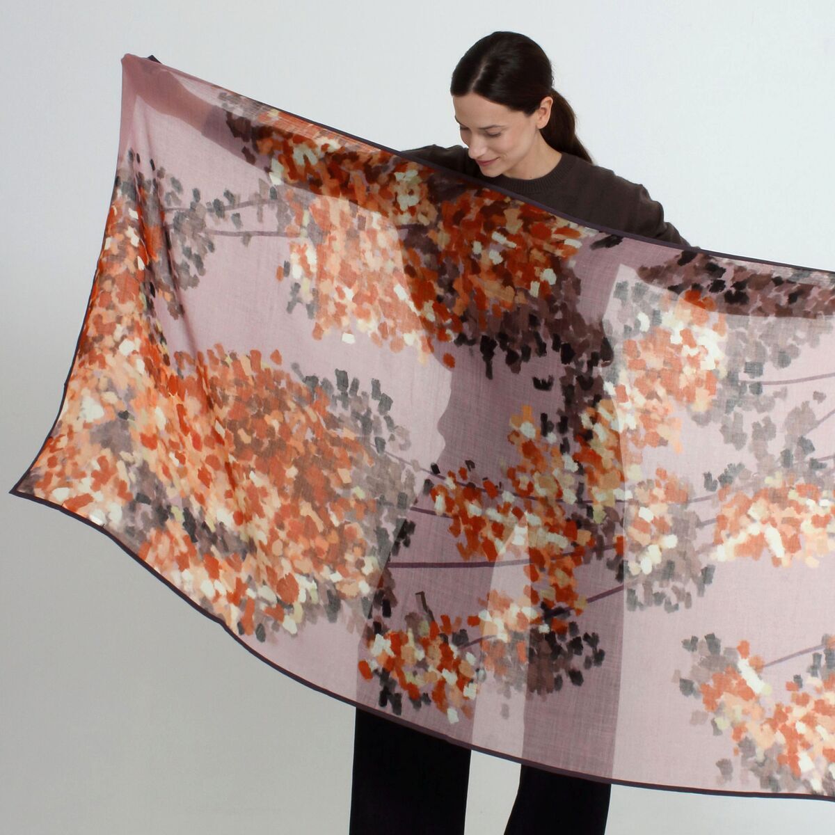 Eine Frau hält den großen Wollschal »Hazel« Mauve von Shalls Barcelona aus 100% Schurwolle ausgebreitet vor sich. Der Schal ist rechteckig, hat ein florales Muster in mauve, orange, creme, grau und schwarz und wirkt weich und stilvoll. Hintergrund neutral hell.
