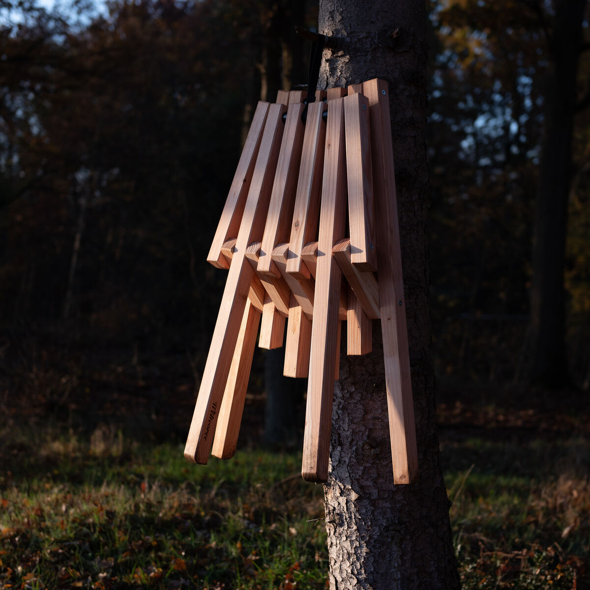 Ein zusammenklappbarer Holzstuhl mit Schafsfell, der an einem Baum im Wald hängt, umgeben von herbstlichen Bäumen und Laub auf dem Boden.
