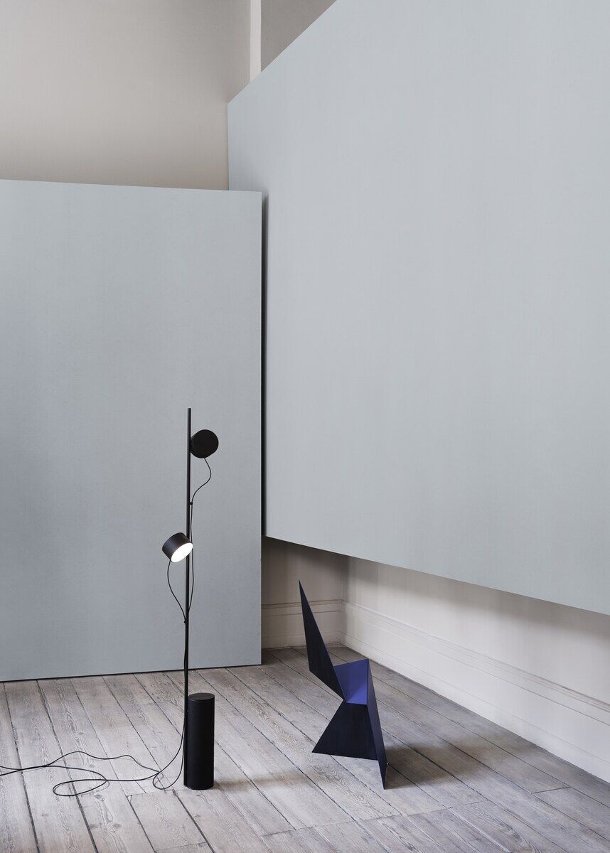 Moderne Bodenlampe »Post« in einem schlicht eingerichteten Raum mit grauen Wänden und Holzfußboden. Die Lampe ist schwarz und besteht aus mehreren kreisförmigen Elementen. Daneben steht ein dunkler geometrischer Stuhl.