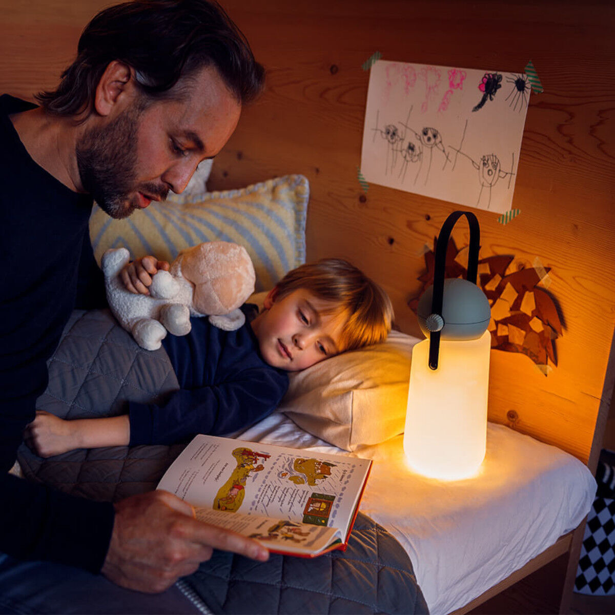 Ein Vater sitzt am Bett seines Kindes, das eine Kuscheltier hält. Neben dem Bett steht eine »Guidelight« Lampe, die warmweißes Licht ausstrahlt. Der Vater liest seinem Kind ein Buch vor, während eine warme und gemütliche Atmosphäre den Raum erfüllt.