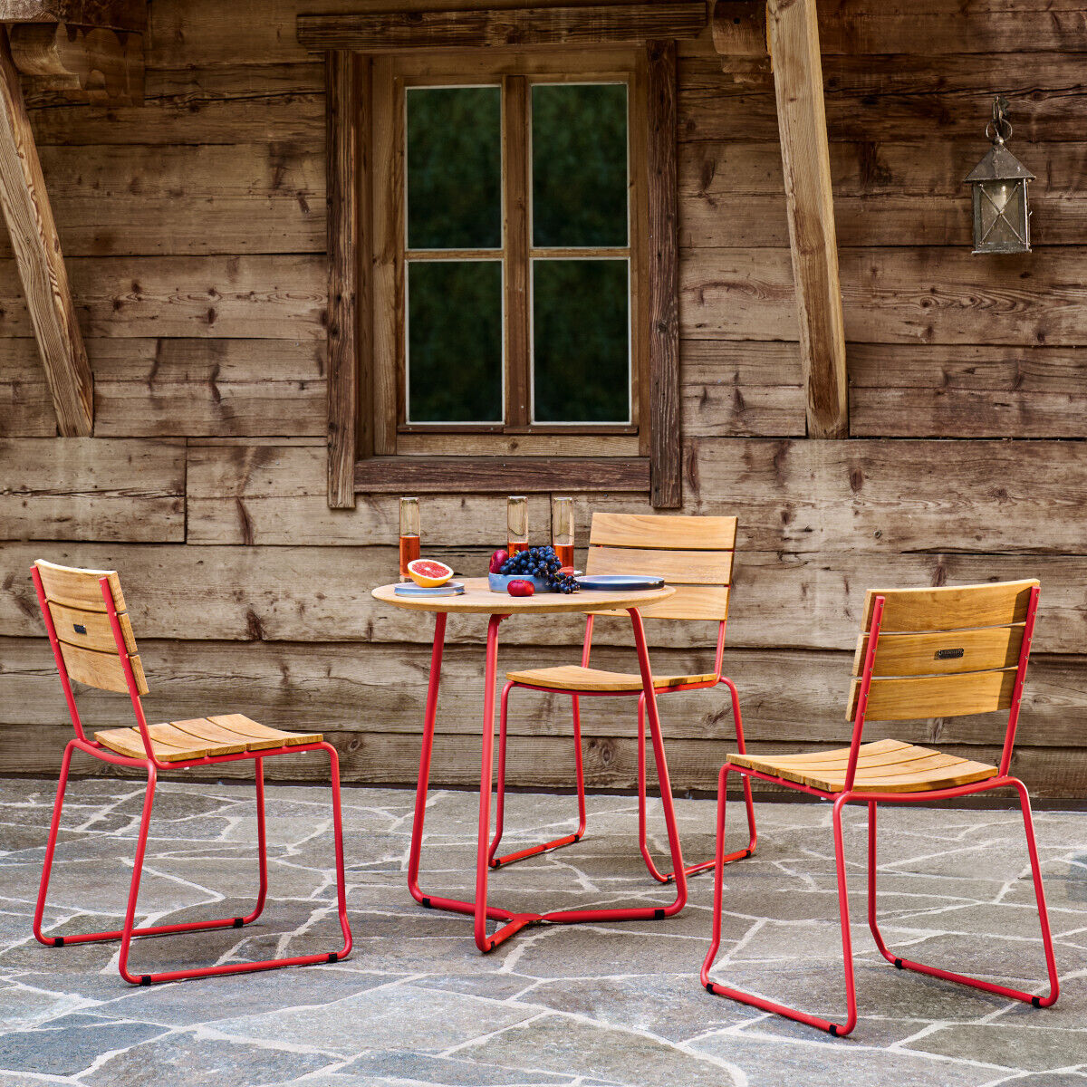 Ein rustikales Outdoor-Sitzset bestehend aus einem Tisch und vier Stühlen aus Teakholz mit roten Metallrahmen steht vor einer holzverkleideten Wand. Das Set ist auf grauen Fliesen platziert.