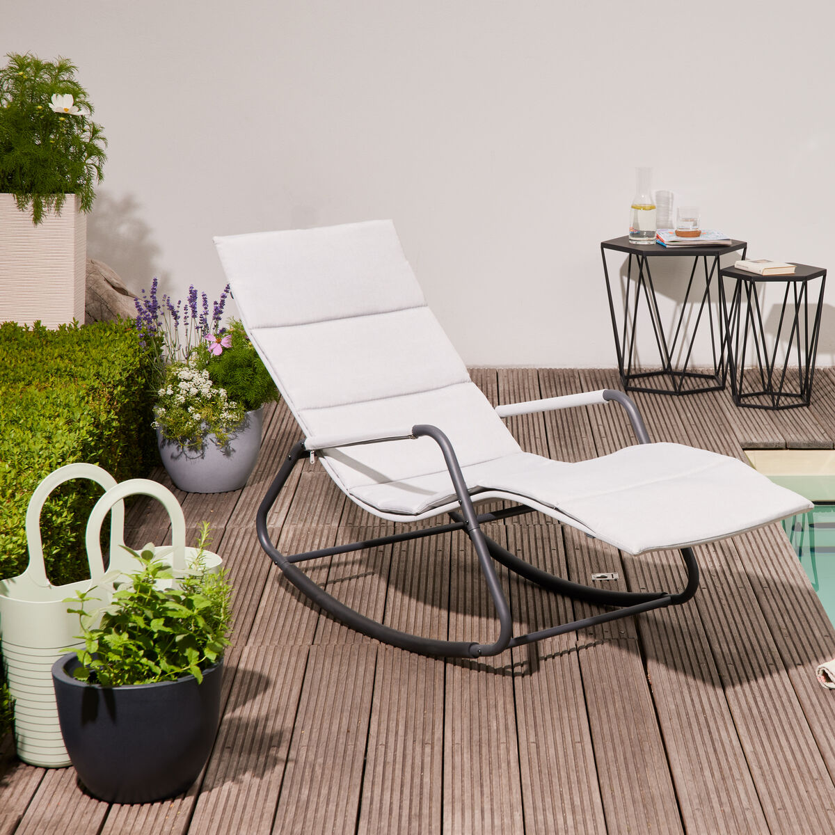 Moderne weiße Swingliege 'Aliano' mit schwarzem Metallrahmen steht auf einer Holzterrasse neben einem Pool, umgeben von Pflanzen in Töpfen und dekorativen Beistelltischen mit Getränken, ideal für entspanntes Sonnenbaden im Garten oder auf der Terrasse.