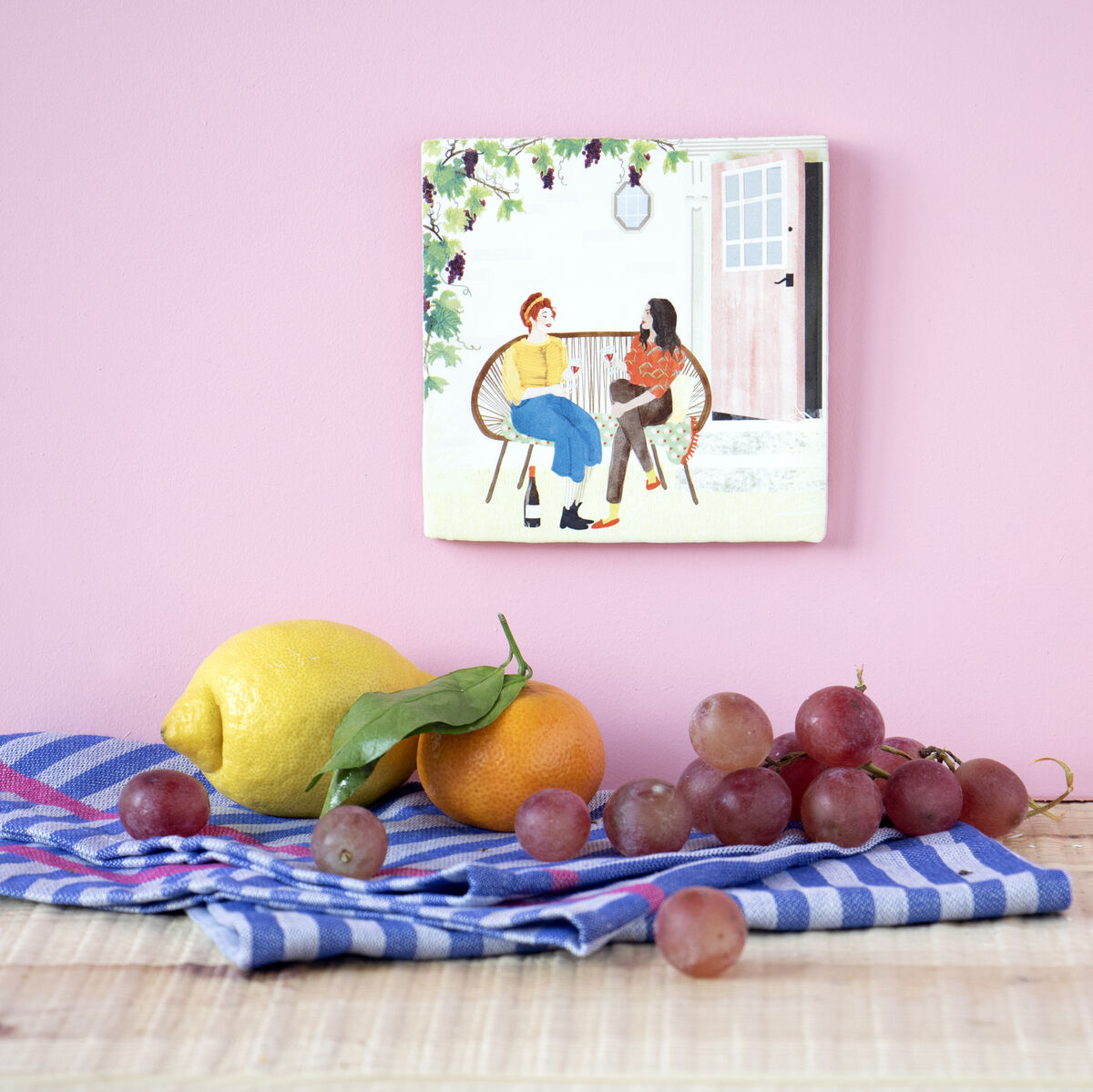 Eine Keramikfliese mit einem Bild von zwei Personen, die Wein trinken, hängt an einer lila Wand über einer Tischdecke mit einer Zitrone, einer Orange und roten Weintrauben.