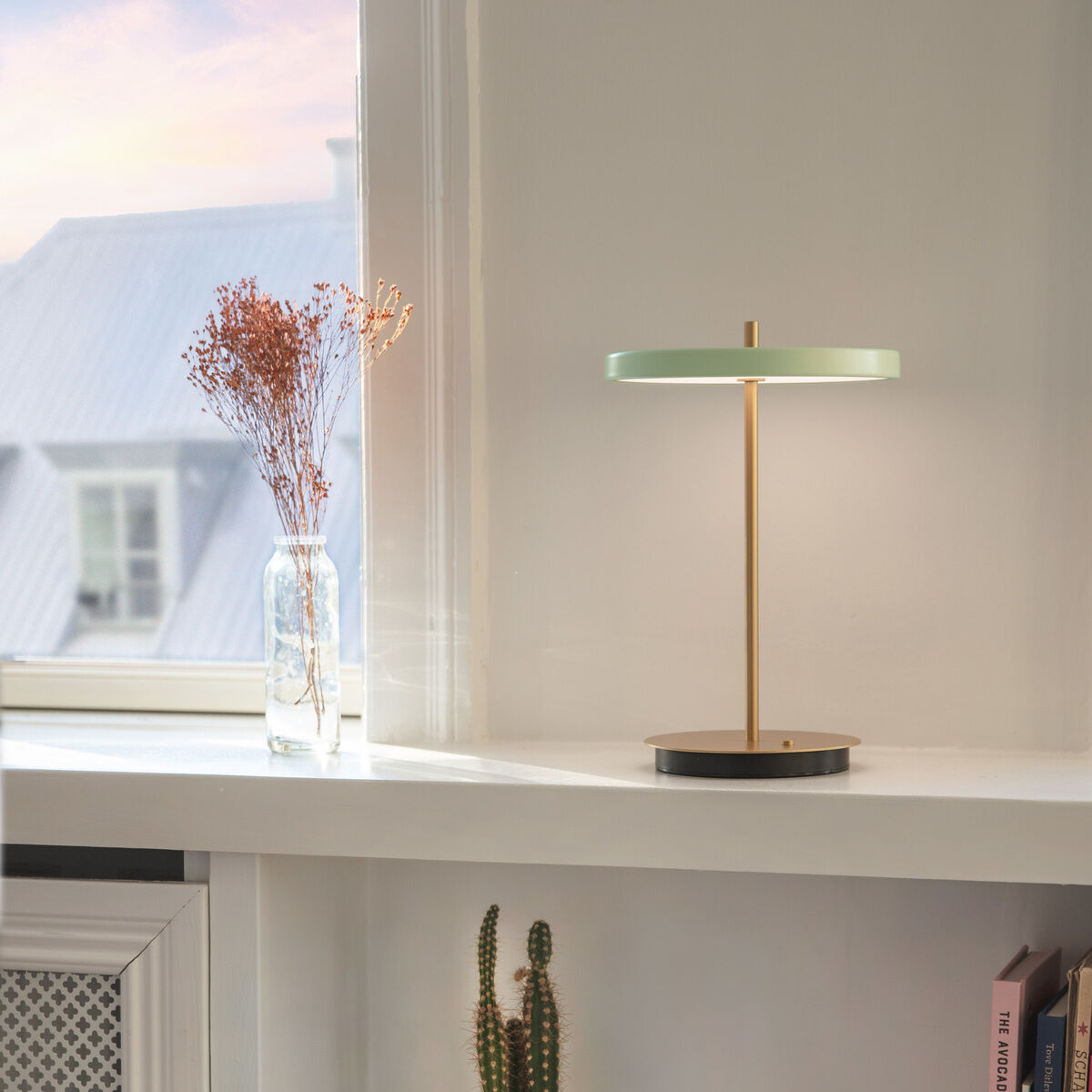 Eine elegante Tischlampe »Asteria Move II« steht auf einer Fensterbank, die von weißen Wänden und einem Bücherregal umgeben ist. Die Lampe hat einen goldenen, schlanken Ständer und einen kreisförmigen, grünen Leuchtkörper. Im Hintergrund sind getrocknete Blumen in einer Vase und ein kleiner Kaktus auf dem Regal sichtbar.