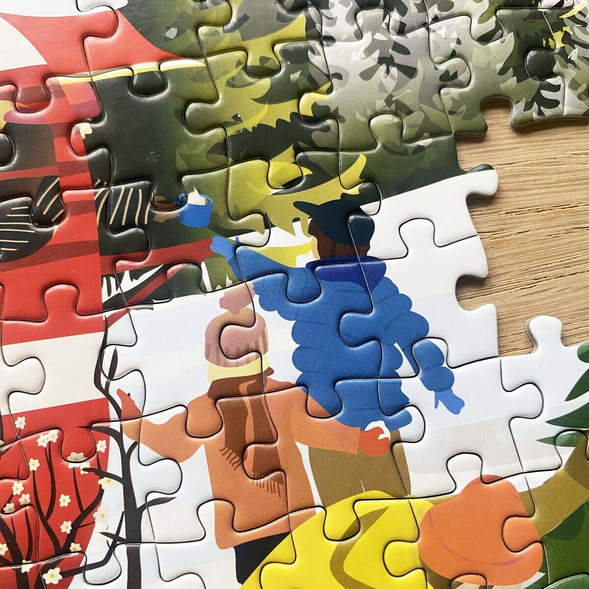 Detailaufnahme des 300 Teile Puzzles »Caravan« von VISSEVASSE. Motiv: stilisierte Figuren in bunter Winterkleidung bei winterlicher Outdoor-Szenerie, darunter Kinder mit Schlitten, Bäumen, Schnee sowie ein rotes Zelt. Puzzleteile teilweise zusammengesetzt.
