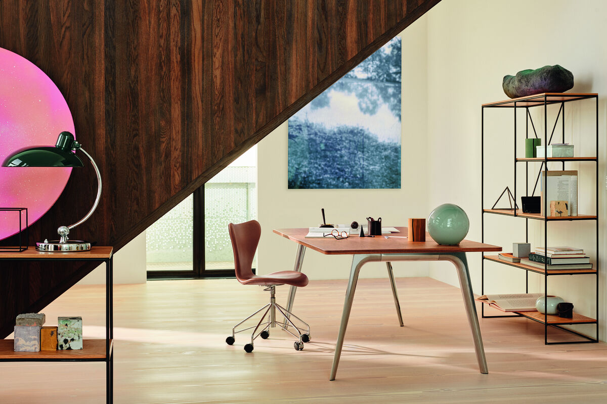 Modernes Bürozimmer mit einem Bürostuhl der Serie 7™. Der Raum ist minimalistisch gestaltet mit Holztönen und Grünakzenten. Ein langes Holzregal, ein Schreibtisch mit hellbrauner Oberfläche und Dekorationsobjekte sind im Hintergrund sichtbar.