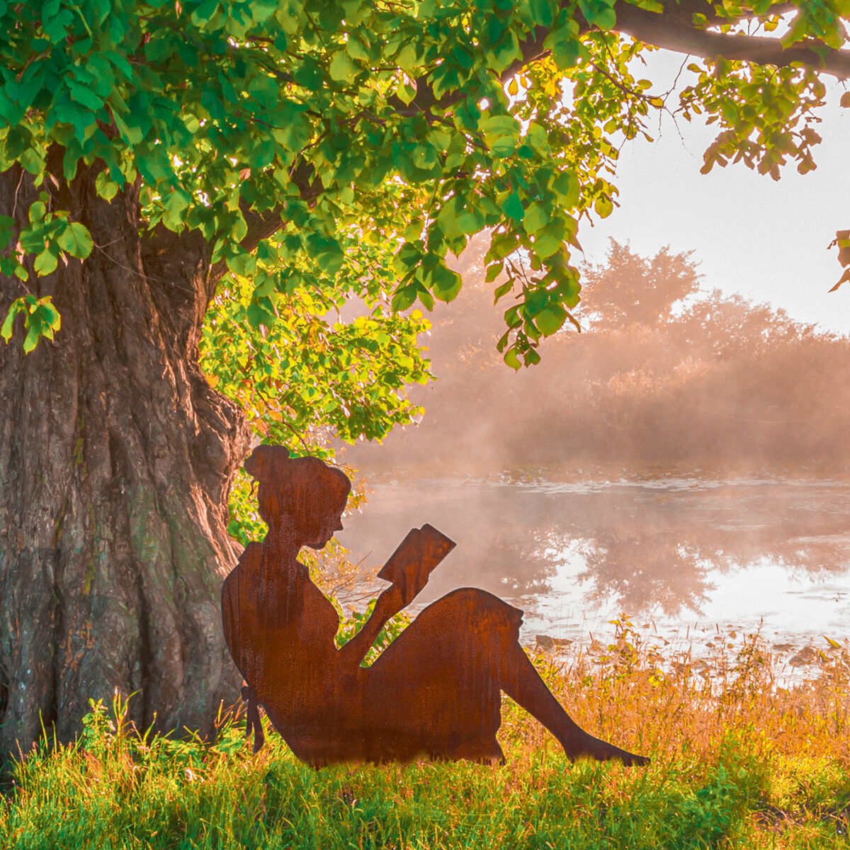 Eine Gartenfigur in Form einer lesenden Silhouette aus Metall vor einem großen Baum und einem See im Hintergrund, umgeben von grüner Vegetation und einem Sonnenaufgang.