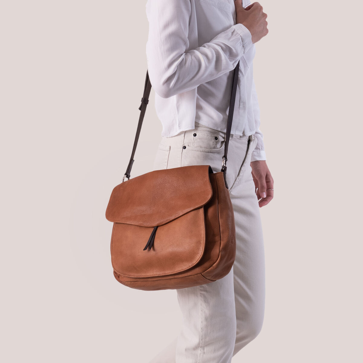 Eine Frau trägt eine braune Leder-Umhängetasche (»Slingbag«). Sie trägt ein weißes Hemd und eine helle Hose. Der Hintergrund ist schlicht und einfarbig gehalten.
