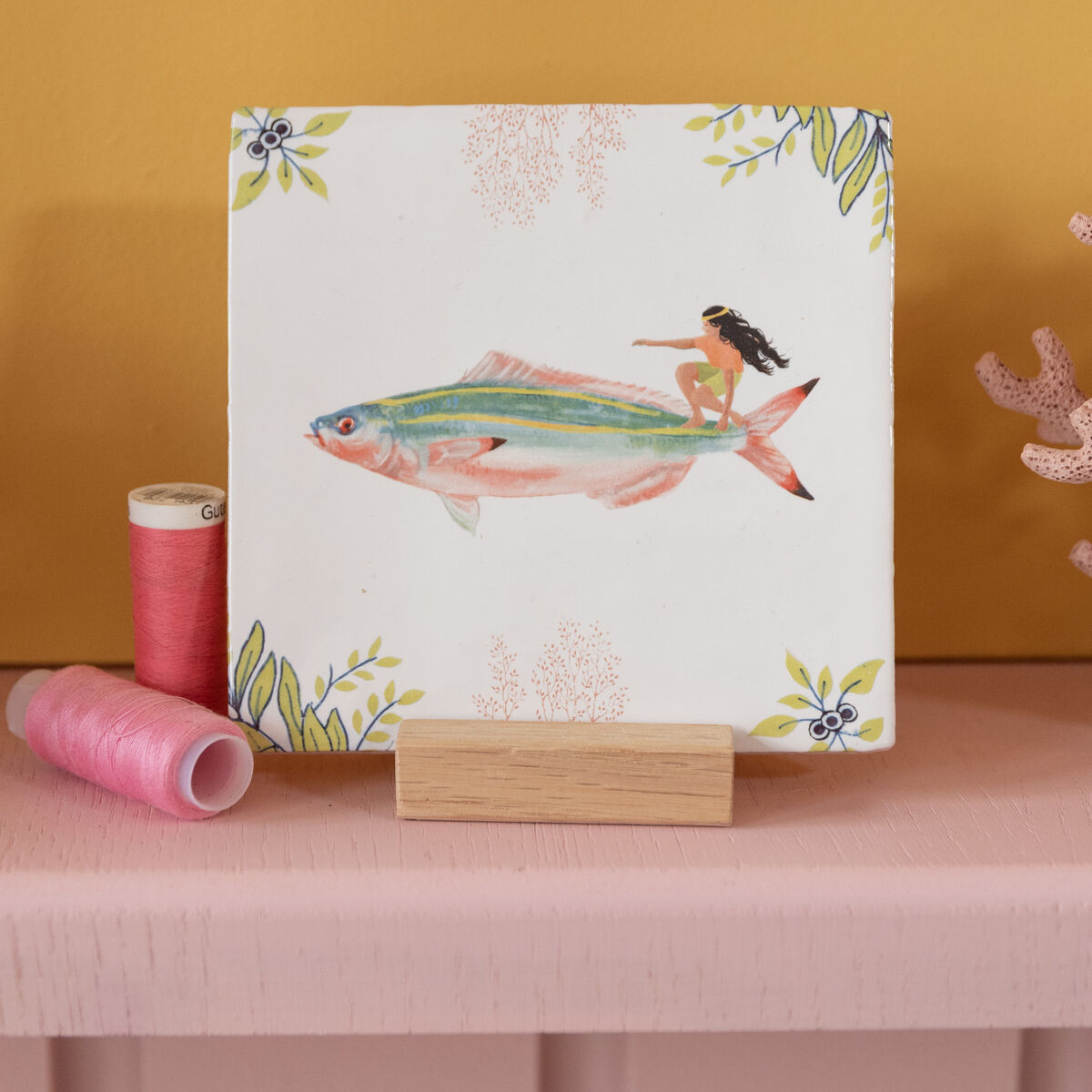 Keramikfliese mit dem Motiv »Auf den Wellen reiten«, auf der eine Person auf einem großen Fisch surft. Die Fliese steht auf einem Regal vor einer gelben Wand, dekoriert mit rosa Garnrollen und einer korallenähnlichen Figur.