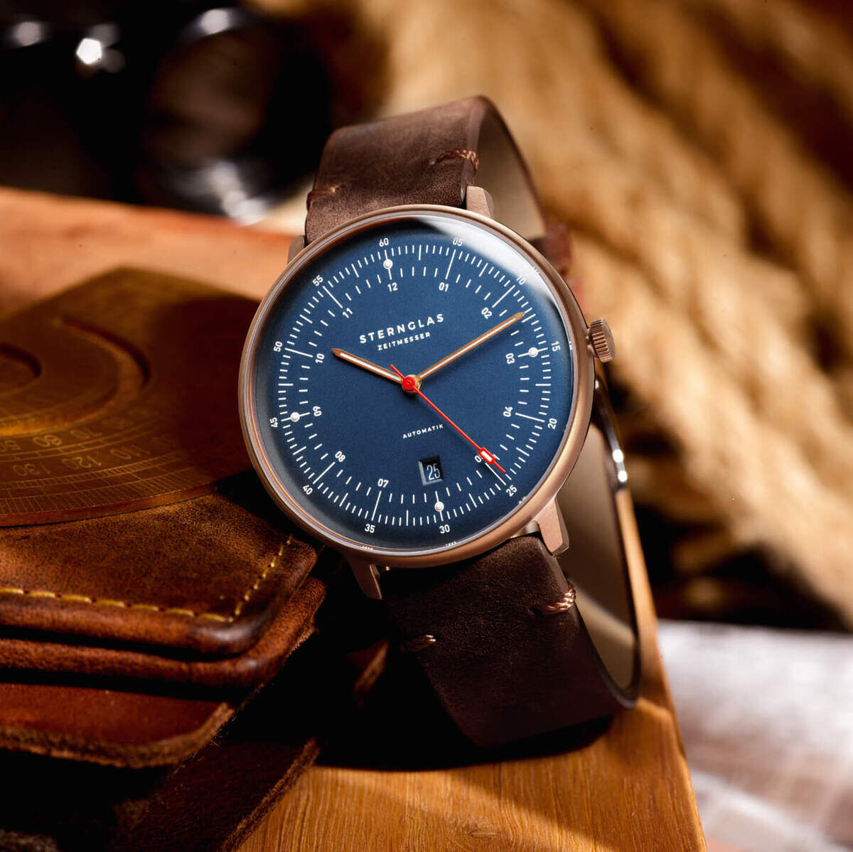 Eine luxuriöse Armbanduhr der ZEIT-Sonderedition namens »Hamburg« mit einem Bronzekorpus. Die Uhr hat ein blaues Zifferblatt mit roten Akzenten und ein braunes Lederarmband. Sie liegt auf einer ledernen Unterlage in einer warmen, gemütlichen Umgebung.