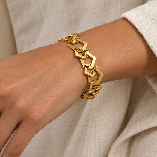 Auf dem Bild ist ein goldfarbenes, geometrisch geformtes Armband aus 22 Karat vergoldetem Messing am Handgelenk einer Person zu sehen. Das Armband gehört zum Schmuckset »Rosa« von Ars mundi und kommt besonders auf heller Kleidung elegant zur Geltung.