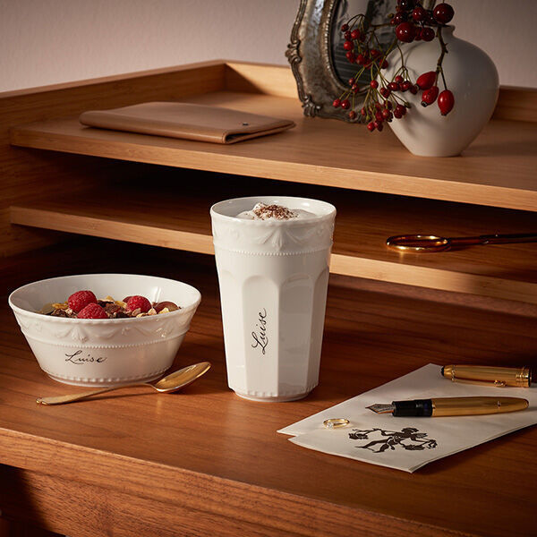 Das Bild zeigt ein Müsli-Gourmet-Set »Kurland« von KPM mit der Schriftart Sententia. Das Set besteht aus einer weiß glasierten Porzellanschale mit einer goldfarbenen Löffel und einer ebenfalls aus Porzellan gefertigten Becher sowie Früchte und Beeren auf einem Holztisch. Im Hintergrund sind weitere dekorative Elemente zu sehen.
