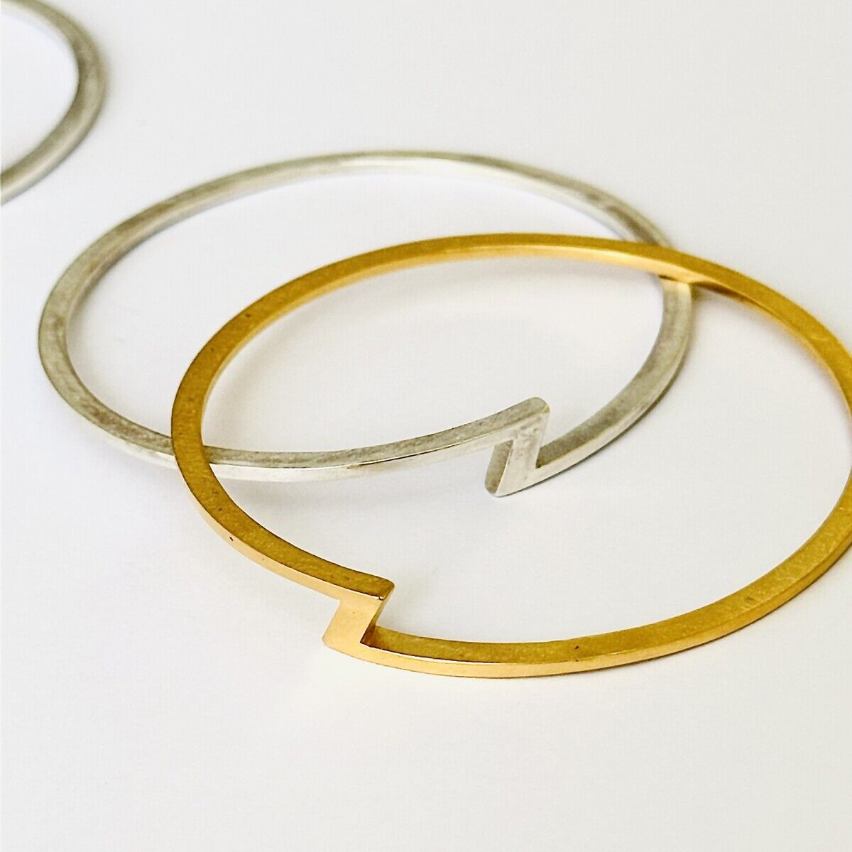 Zwei filigrane Armreife auf weißem Hintergrund, einer feingoldplattiert und der andere aus Silber. Beide zeigen ein markantes Z-förmiges Designelement. Die Armreifen wirken modern und hochwertig, geeignet als elegantes Schmuckstück oder Geschenk.