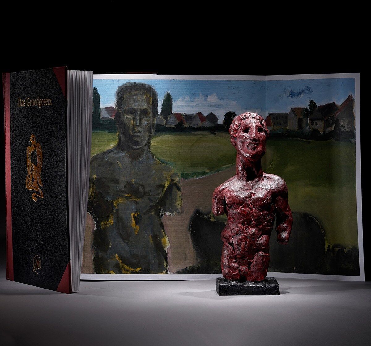 Das Bild zeigt eine Skulptur von Markus Lüpertz mit dem Titel »Das Grundgesetz« aus dem Jahr 2013. Die Skulptur ist rot und steht vor einem aufgeschlagenen Buch, das ebenfalls das Grundgesetz darstellt. Im Hintergrund befindet sich ein geteiltes Gemälde mit zwei menschlichen Figuren und einer Landschaft mit Häusern.