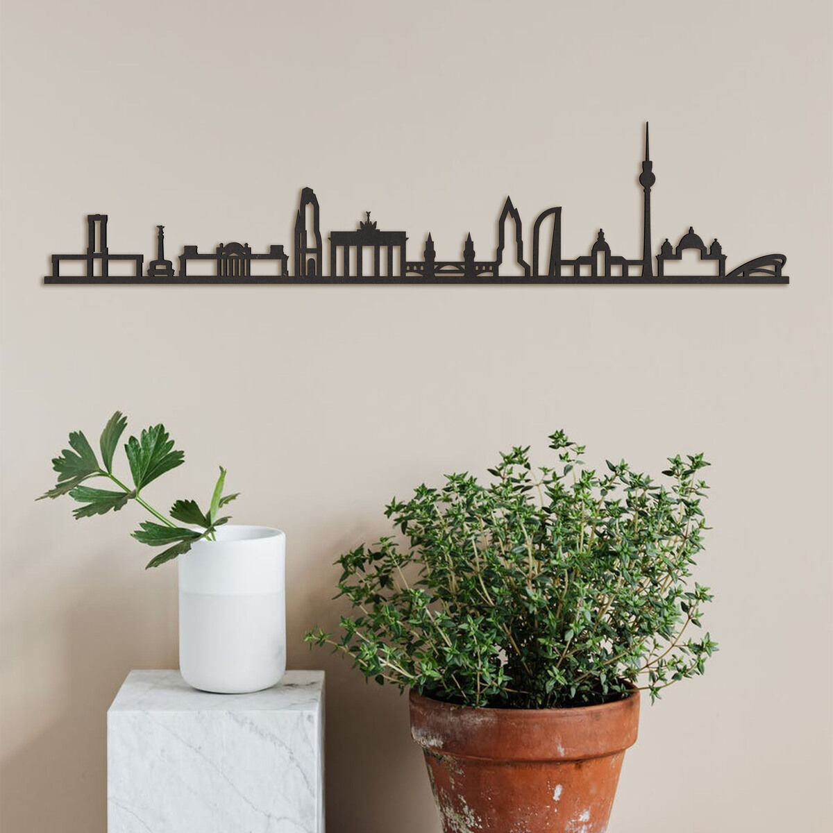 Holz-Skyline von Berlin an einer beigen Wand montiert, neben einem weißen Sockel mit einer grünen Pflanze darin und einem großen Terrakottatopf mit einer weiteren grünen Pflanze.