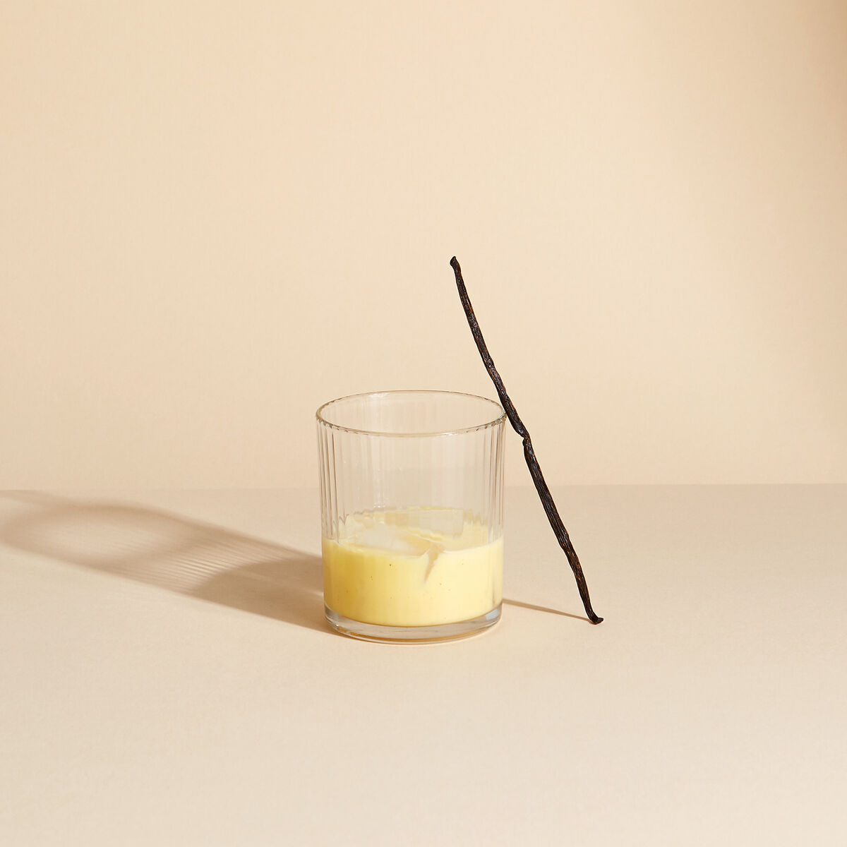 Ein Glas mit gelbem Eierlikör und einer Vanilleschote, das auf einer beigen Oberfläche steht.