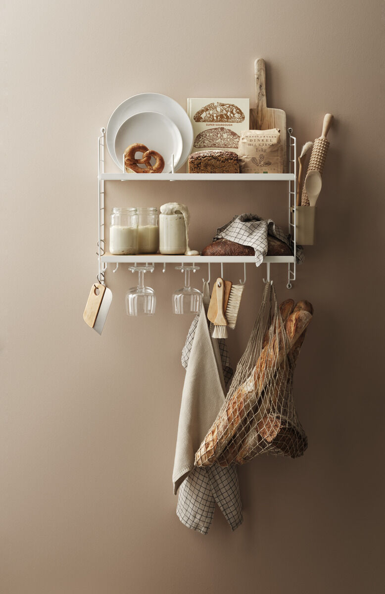 Das Bild zeigt ein weißes, modernes Küchenregal von String an einer beige gestrichenen Wand. Auf dem Regal stehen Schüsseln, Bücher, ein Holzbrett und Gläser. Zudem hängen Geschirrtücher und ein Netz mit Brot am Regal.