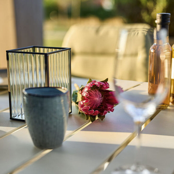 Ein Tischmodell 'VAPIO' mit den Maßen 180x90 cm steht in einem Essbereich im Freien. Auf dem Tisch befinden sich dekorative Elemente wie Kerzen, eine Laterne, zwei Weinflaschen und eine Blume. Der Hintergrund zeigt eine grüne Landschaft, die verschwommen ist.