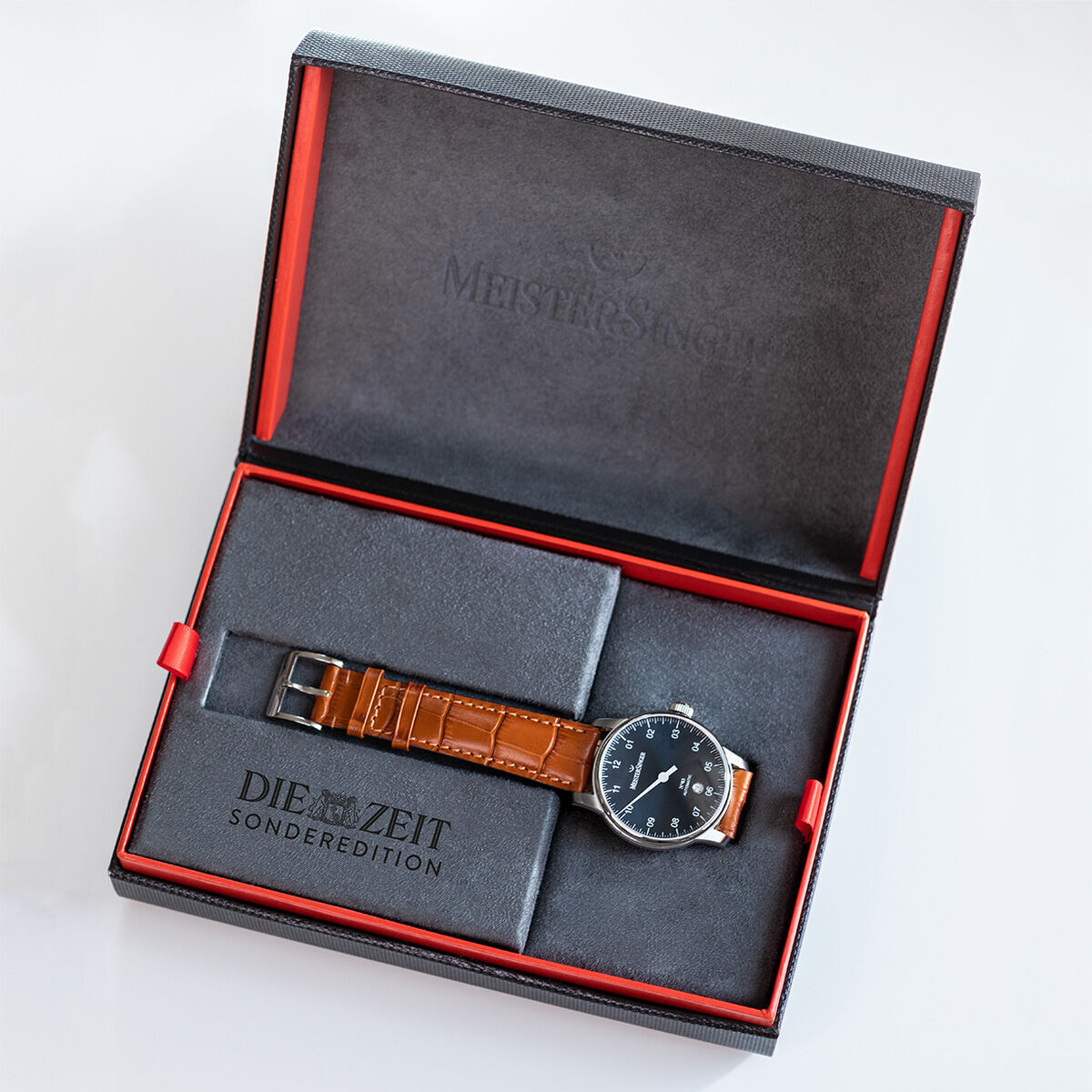 Eine ZEIT-Sonderedition »N°03« Armbanduhr von MeisterSinger mit einem braunen Lederarmband und einem schwarzen Zifferblatt, präsentiert in einer eleganten schwarzen Box mit rotem Innenfutter und einem Fach für die Uhr.