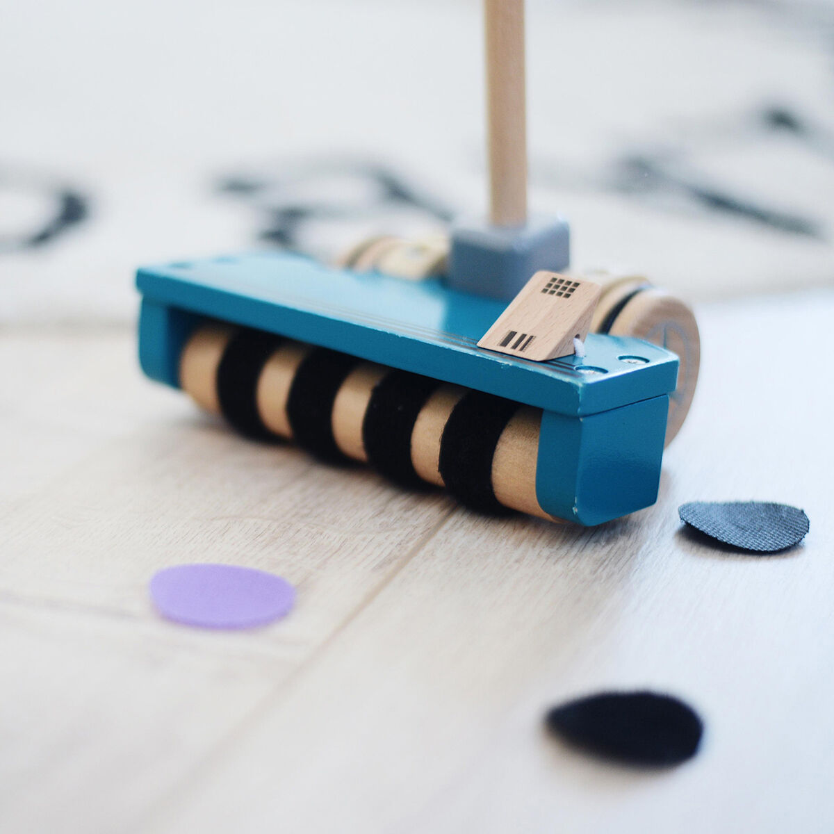 Ein buntes Schiebespielzeug in Form eines Staubsaugers auf einem hellen Holzboden, mit bunten Filzscheiben in Lila und Schwarz herumliegend.