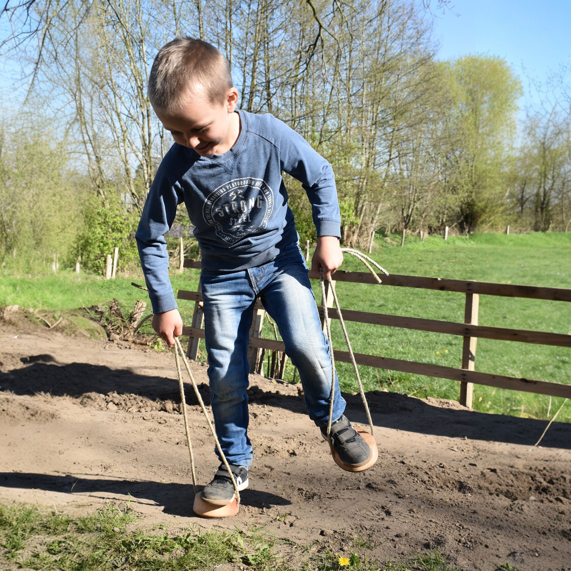 Ein Junge spielt draußen auf einem sandigen Weg mit Holzstelzen, die echte Hufeisen an der Unterseite haben. Die Stelzen werden mit Griffseilen aus Hanf gehalten. Im Hintergrund sind ein Holzzaun, grüne Wiese und lichte Bäume unter blauem Himmel zu sehen.